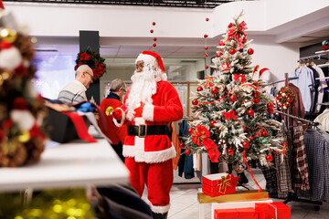 Fototapeta premium Employee Santa Claus in Christmas themed store, jingling xmas bells to attract customers in shopping center. Retail assistant dressed in festive costume greeting clients in decorated fashion boutique