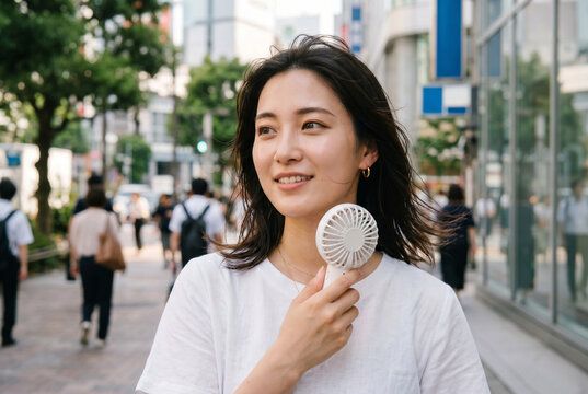 真夏の屋外でハンディファンを使い涼む笑顔の日本人女性。暑さ対策、熱中症予防。Generative AI