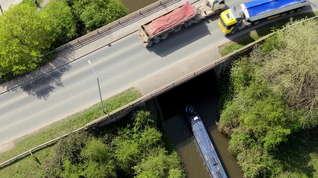 Aerial view of a narrowboat cruising along a canal and passing under a road bridge surrounded by lush green trees in Barton-under-Needwood, England, United Kingdom.