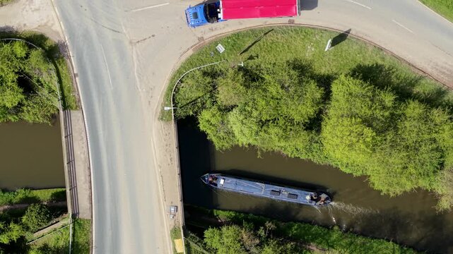 Aerial view of a narrowboat cruising along a canal under a road bridge with lush green trees and a truck in Barton-under-Needwood, England, United Kingdom.