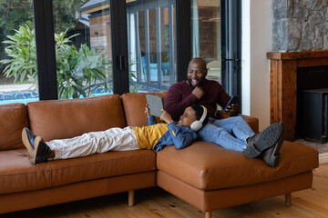 Obraz premium African American father and son lounging at home, son using tablet while father holding phone