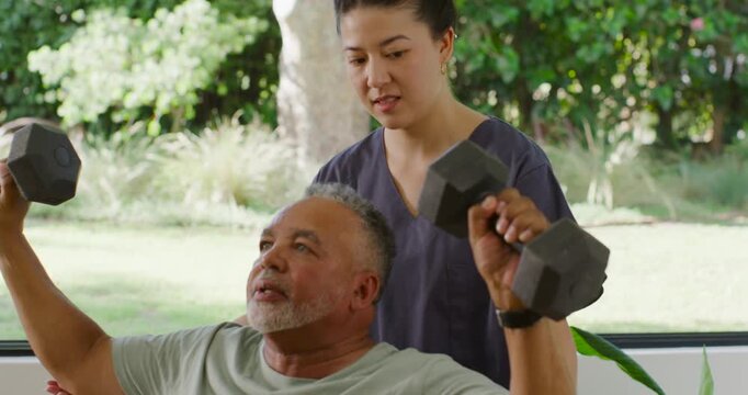 Senior man pressing dumbbells on bench on PT cue, Asian caregiver in scrubs spotting for rehab