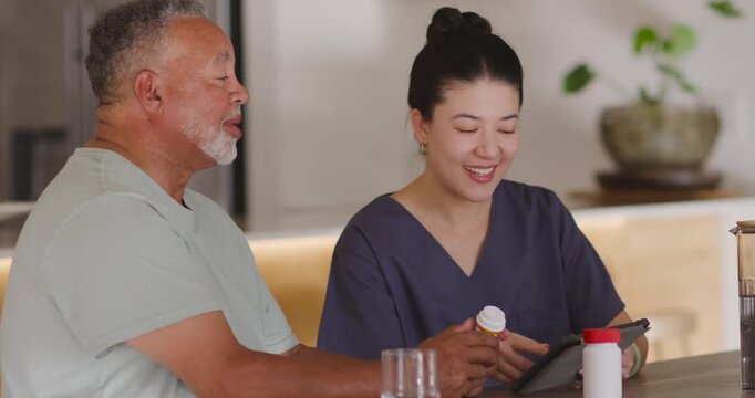 Asian caregiver in scrubs guiding man holding yellow bottle, checking dose on tablet at home