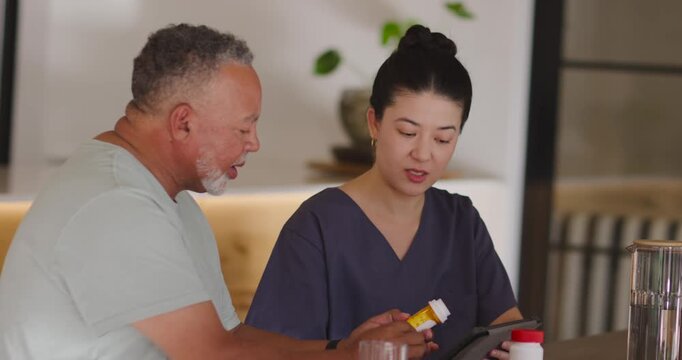 Asian caregiver holding yellow bottle and Senior man clarifying dosage using tablet at home table