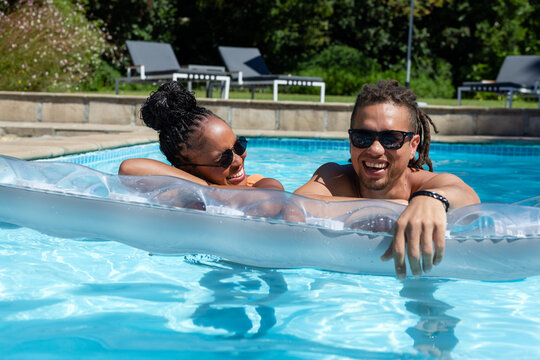 African American couple lounging on clear inflatable float in swimwear in pool, wearing sunglasses