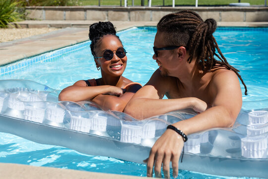 African American couple leaning on clear float in backyard pool, wearing swimwear and sunglasses