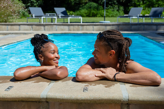 African American couple leaning on pool edge, smiling at each other in swimwear with sunglasses