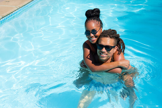 African American couple in shades and swimwear riding piggyback and splashing in sunny outdoor pool