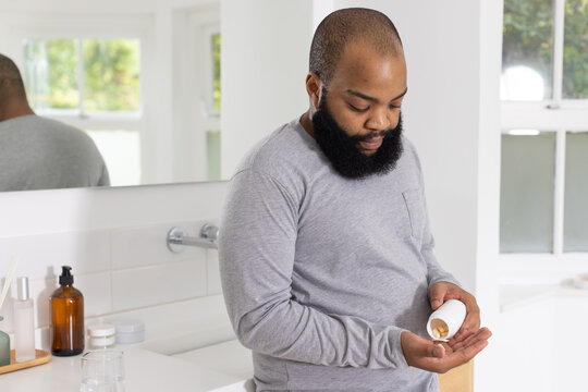 African American man standing at bathroom sink pouring tan pills from white bottle with beard