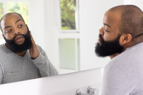 African man standing at bathroom sink checking and touching beard in wall mirror near chrome faucet