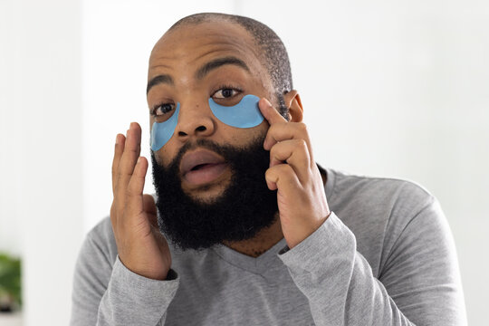 African American man applying blue hydrogel under-eye patches at vanity, wearing gray shirt