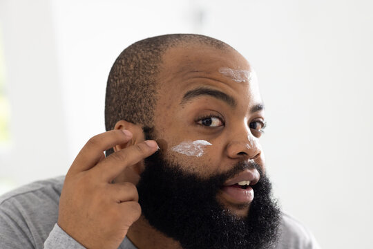 Adult African American man applying white cream to cheek with fingers in bathroom, wearing gray top