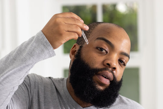 African man applying dropper to beard at white-framed window, wearing light gray long-sleeve shirt