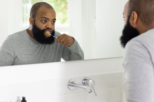African man brushing teeth at bathroom sink and mirror, holding multicolor toothbrush in gray shirt