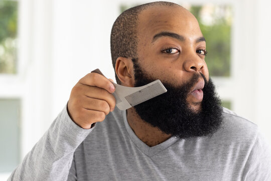 African adult male grooming thick beard, holding light-gray comb near large window, in gray V-neck