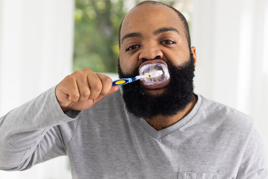 African man brushing teeth at sink near window with blue toothbrush and clear tray, gray shirt