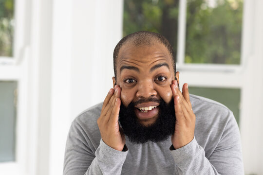 African man pressing palms to cheeks, smiling by window panes, wearing light gray shirt, full beard