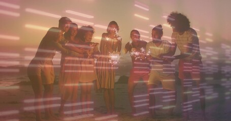 Fototapeta premium Waving sparklers, eight adults in summer clothes posing on beach at sunset, with streaked light
