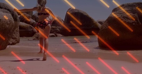 Fototapeta premium Wading girl in star bikini moving into shallow sea on beach large boulders neon-orange or pink strea