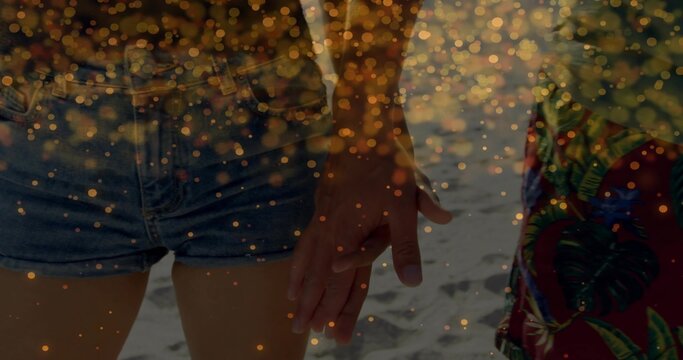 Holding hands couple standing on sand, wearing denim cutoff shorts, colorful garment, golden bokeh