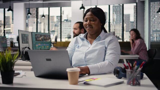Black female staff working on laptop at desk in modern corporate workspace, ensuring business administration for strategy development. Businesswoman ensuring productivity in open space.