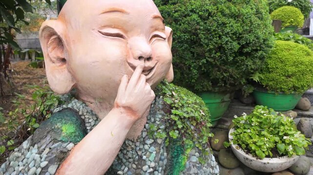 A smiling stone statue of a child, adorned with moss and pebbles, sits in a lush garden in Ba Na Hills, Da Nang, Vietnam. The statue evokes a sense of peace and joy.