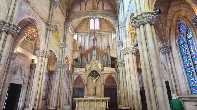 A man in a green jacket and hat walks through the interior of a Catholic church in Ba Na Hills, Da Nang, Vietnam, past rows of wooden pews.