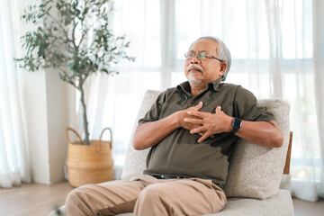 Fototapeta premium Elderly health insurance, elderly man chest pain sitting on chair at home, eyes closed and hand on chest with discomfort