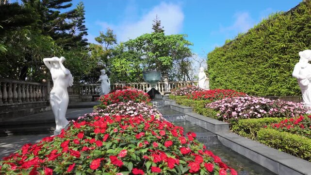 A beautiful garden in Ba Na Hills, Da Nang, Vietnam, features white statues amidst vibrant red and pink flowers, creating a serene and picturesque landscape.