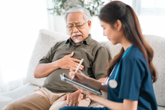 Caregiver with Asian senior patient, senior man with chest pain during home health checkup by nurse, worried patient receiving care in living room