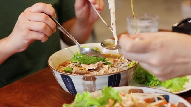 Close up video of hands of two people eating a delicious bowl of vietnamese pho soup noodles with fresh vegetables and meat at a restaurant.