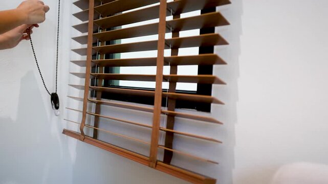 Close up of person hand pulling cord to adjust brown wooden window blinds in bright room.
