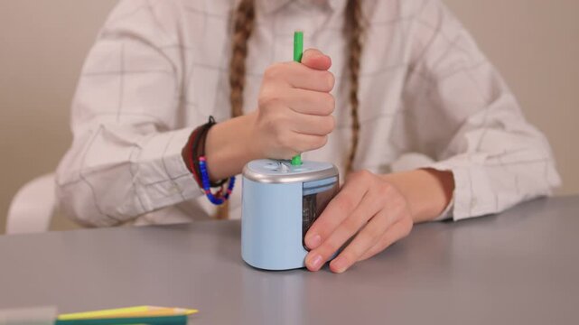 Hands rotating crank to sharpen. Focused student sharpening pencil with colorful accessories visible. Student attentively sharpening green pencil using hand crank on desk