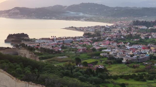 erial view of the peaceful coastal resort town of Sidari in Corfu, Greece at dawn, showcasing the beautiful sandy beach, calm Ionian sea, traditional houses, and lush green hillsides.