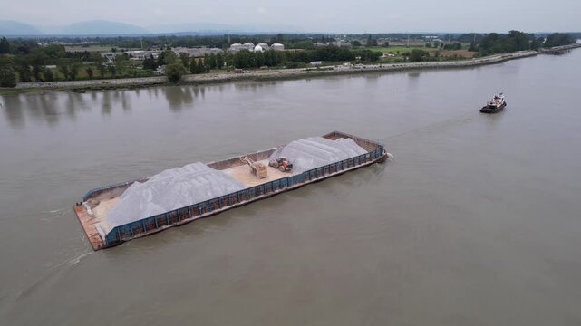 Small tugboat pulling loaded gravel barge up river with a frontend loader.