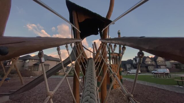 Thick woven climbing rope stretching across timber play structure hanging above wood chips framed against suburban housing beneath cloudy sunset sky captured via backward moving viewpoint