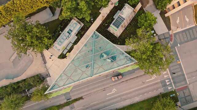 Triangular architectural structure sheltering smart hydrogen refueling equipment bordered by campus roadway and concrete skatepark under bright daylight captured via slow rotating top down aerial pers