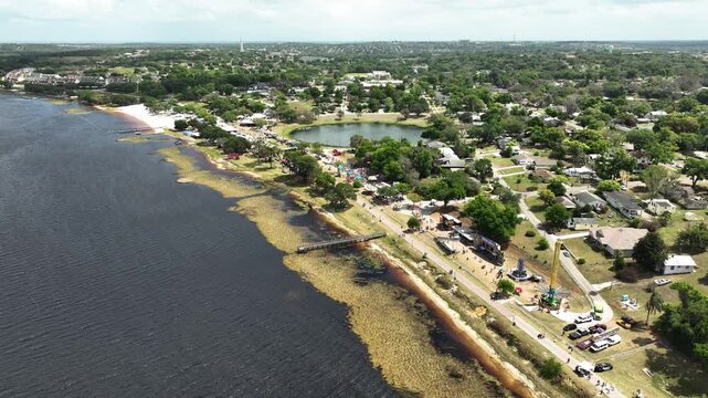 A cinematic drone view captures the Pig on the Pond festival in Clermont, Florida, set along the scenic waterfront with crowds, rides, and community gatherings.