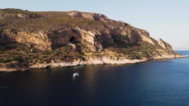 Aerial drone shot of dramatic limestone cliffs with a sea cave along the Favignana coastline, Sicily. A white boat drifts on clear Mediterranean waters below.