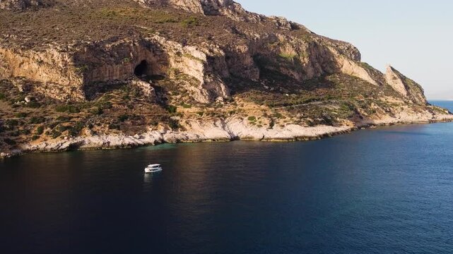Stunning aerial drone footage of a rugged limestone coastal cliff in Sicily, Italy, featuring a natural sea cave carved into the rock face. A single white boat drifts on crystal-clear turquoise Medite