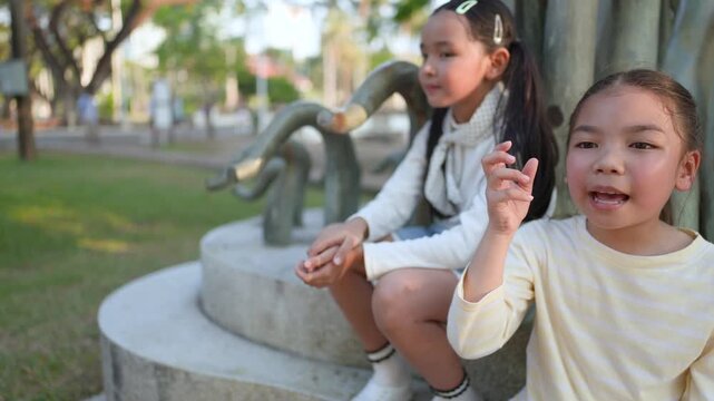 Two Young Asian Sisters Sitting and Talking in a Sunny Bangkok Park