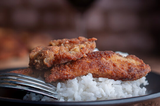 Milanesas de pollo pescado o carne de res sobre arroz blanco al vapor con un tenedor al lado, sobre un plato negro y pared de ladrillos al fondo, macro o acercamiento.