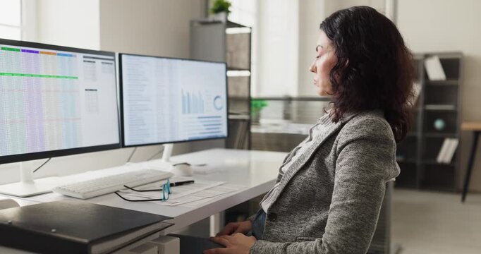 Tired woman analyst in office with monitors. She reviews spreadsheets on dual computer screens, pausing as overwork and fatigue build during a day. Clear workplace stress overload concept.
