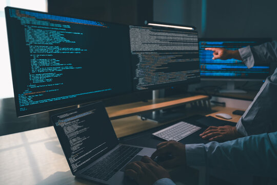 man programmer writing program code at a multi monitor workstation with laptop and software code on screens, performing debugging, software development, and technical workflow in a dark office environ