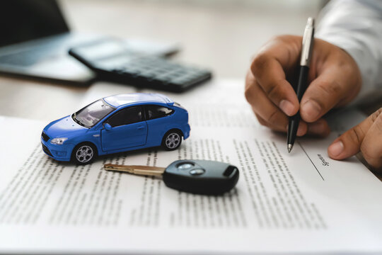 Hand holding pen signing car loan finance document contract with blue toy car model and key on paperwork, representing auto finance agreement approval, vehicle lease purchase, loan application process
