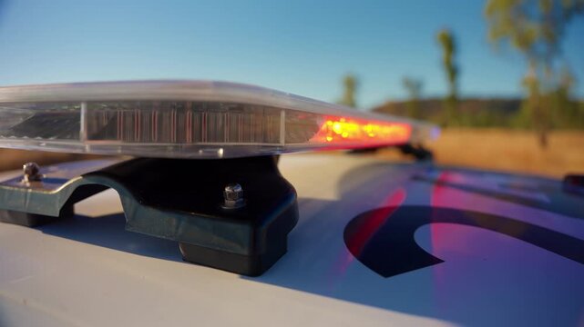 Close-up view of police car light bar with flashing red and blue emergency lights on white patrol vehicle roof against clear blue sky in rural outdoor setting with trees and golden grass.