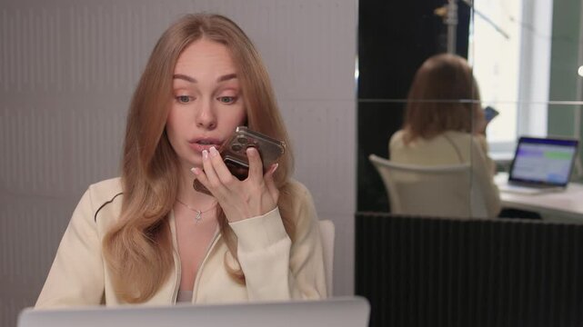 Female candidate communicates via phone with colleague visible in reflection. Caucasian woman engaged in professional remote interview with colleague reflected in glass