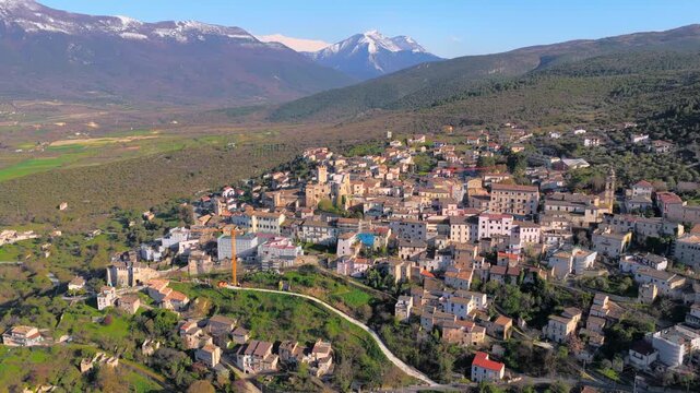 Aerial view of the historic hillside town of Capestrano with stone buildings and lush green valley under snow-capped mountains in Capestrano, Abruzzo, Italy.