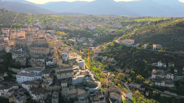 Aerial view of the medieval Castello Piccolomini and traditional stone architecture nestled among lush green hills and mountains in Capestrano, Abruzzo, Italy.