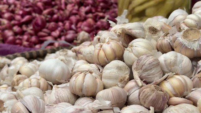 Dense layers of large garlic bulbs cover the selling surface, with stacks of red onions and retail activity visible in the market setting. Food distribution.
